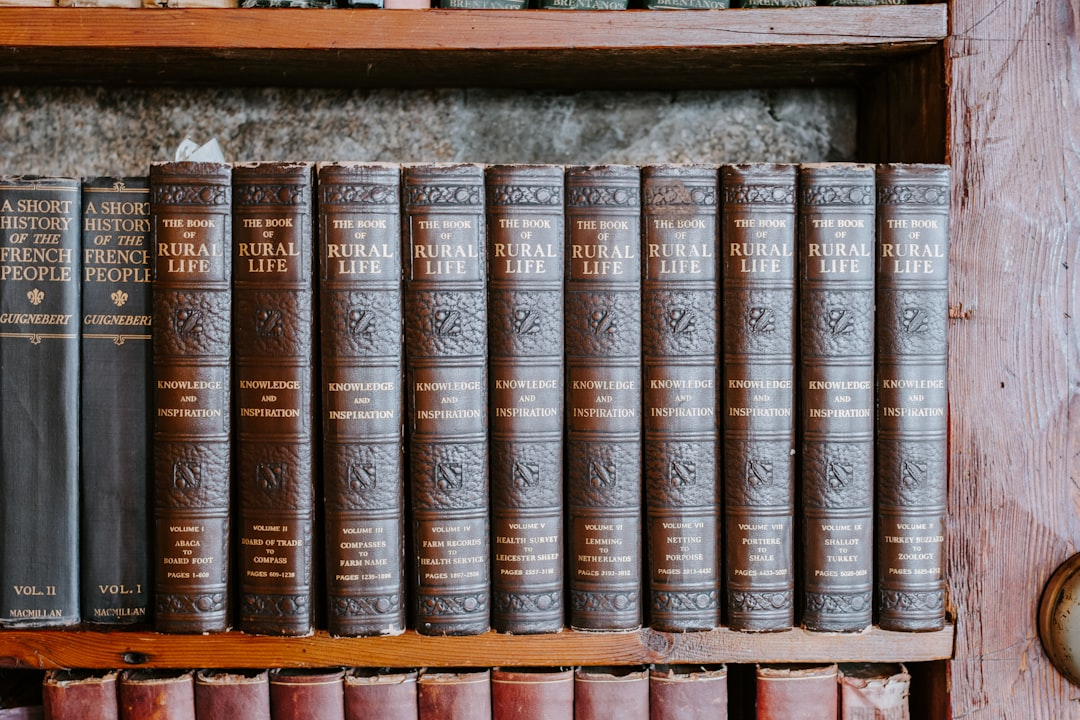 A hand gently placing a glowing, open book onto a neatly organized, substantial stack of diverse books and digital tablets, under soft, inviting light.