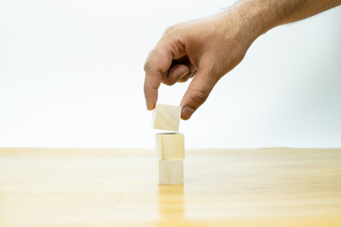 A hand carefully places a third wooden block atop two others on a light surface, symbolizing the progressive and foundational stages of Maslow's Hierarchy of Needs in humanistic psychology.
