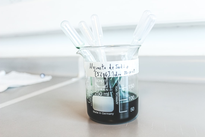 A clear laboratory beaker, labeled 'Alginato de Sodio' and holding a dark liquid with multiple pipettes, sits on a bright counter, representing biotech education and scientific innovation.