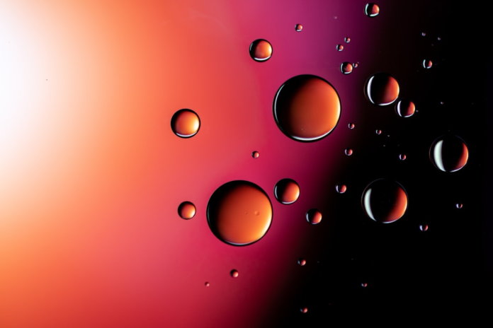 Luminous, multi-sized spheres reflecting warm orange and red light float against a vibrant gradient backdrop of bright white, deep red, and dark purple, evoking an abstract cosmic or scientific realm.