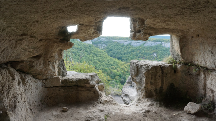 Inside an ancient rock-cut cave, a large, rugged opening frames a panoramic view of a lush green valley and distant forested cliffs under a bright sky, conveying the timeless beauty of historical sites.
