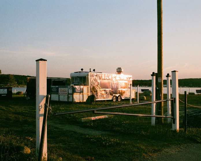 A gleaming silver food truck, featuring 'Holy Gyro' and 'Coming Soon' signs, reflects golden hour light beside a tranquil lake, framed by a grassy foreground and a partially open barrier gate.