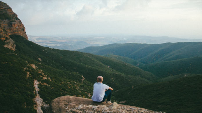 A person sits mindfully on a rocky mountain perch, overlooking expansive green valleys and distant peaks under a hazy sky, embodying serene contemplation and mindfulness practice.