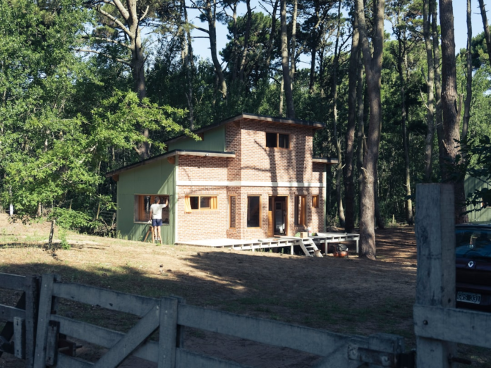 A two-story home, blending green siding and brick, is situated on a sloped grassy clearing within a dense, tall forest, where a person on a ladder performs work, illustrating a residence built with consideration for its natural environment.