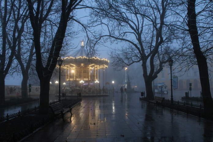 A brightly lit, ornate carousel stands as a focal point in a misty, wet park at dusk, surrounded by the silhouettes of bare trees, creating an enduring and mysterious atmosphere.
