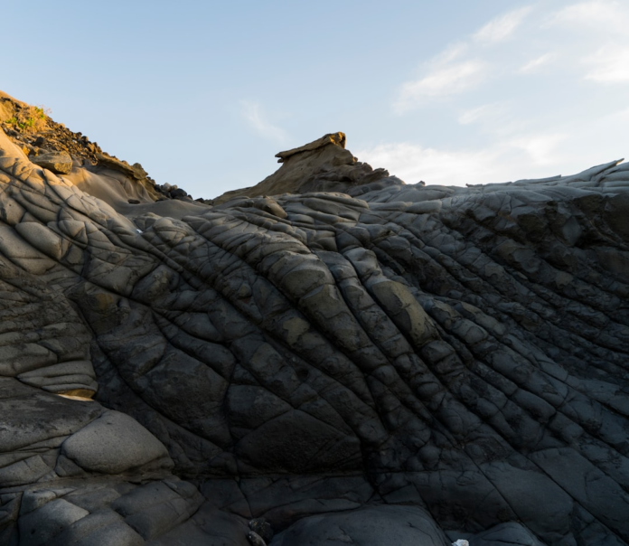 Textured, dark gray geological formations with dramatic folds and a sharp pinnacle reaching towards a pale sky, resembling an alien landscape of extraterrestrial geological wonders.