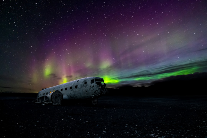 A derelict airplane fuselage rests on a dark, desolate plain beneath a dramatic display of vibrant green and purple Aurora Borealis streaking across a star-filled night sky.