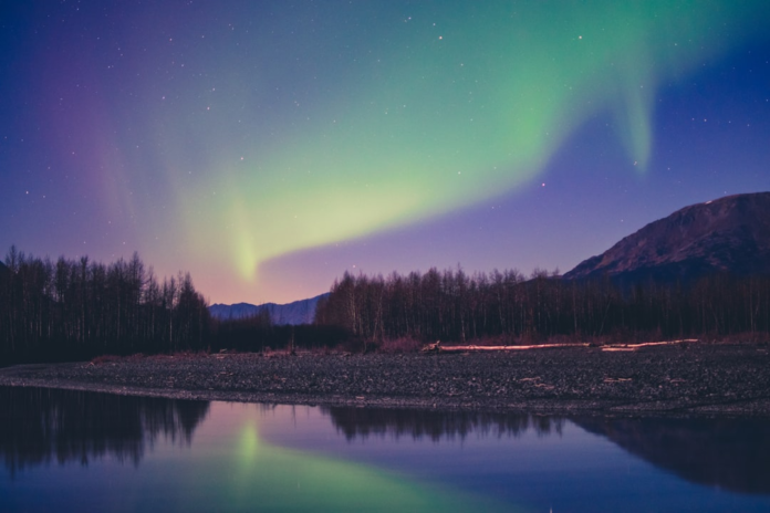 Dazzling green and purple aurora borealis illuminates a starry night sky, reflecting vibrantly on a calm river bordering a dark forest with distant mountains.