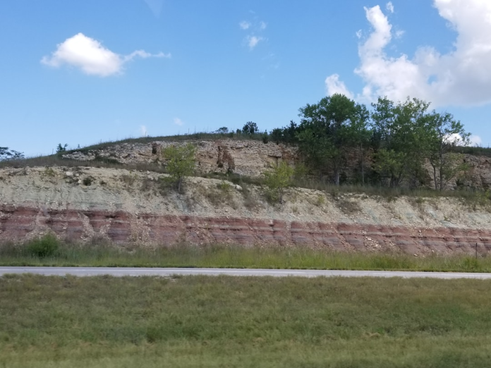 Exposed geological strata reveal distinct reddish-brown and lighter grey rock layers on a hillside beside a road, illustrating the earth's foundational bedrock under a clear blue sky.