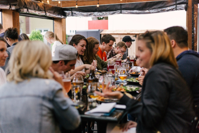 A diverse group of young adults share a lively meal and drinks at a long table on an outdoor patio, illustrating social context and community engagement.