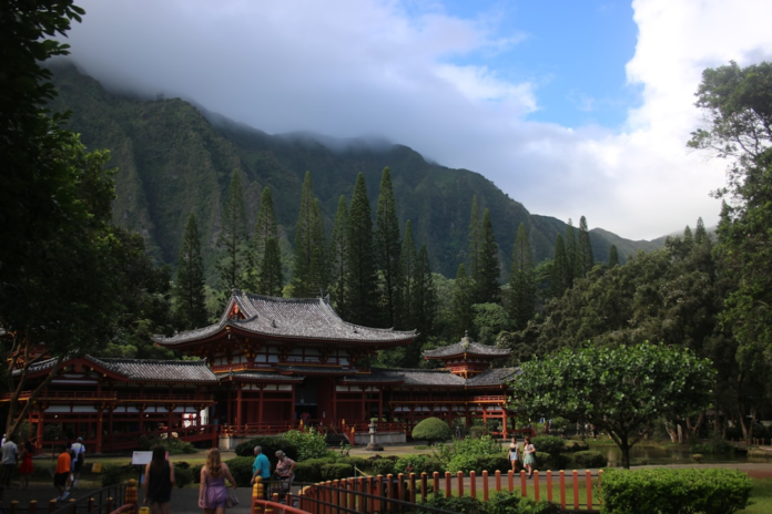 A traditional red and grey Japanese temple complex is nestled amidst a vibrant, diverse landscape of towering evergreen trees and lush, mist-covered mountains under a partly cloudy sky.