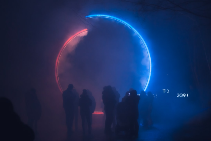 A large, conceptual glowing red and blue ring, resembling a black hole's event horizon, stands prominently in a mysterious, smoky dark space, observed by silhouetted figures.