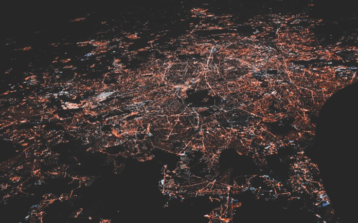 Expansive aerial view of a vibrant city at night, illuminated by a dense, interconnected network of glowing orange and white lights, representing the vast web of smart devices and IoT connectivity.