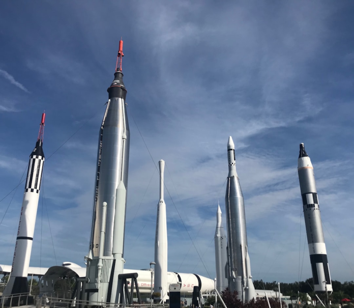 Multiple towering silver and white historical rockets, including Mercury-era spacecraft, stand reaching towards a bright blue, partly cloudy sky, embodying the enduring legacy of space exploration and human ingenuity.