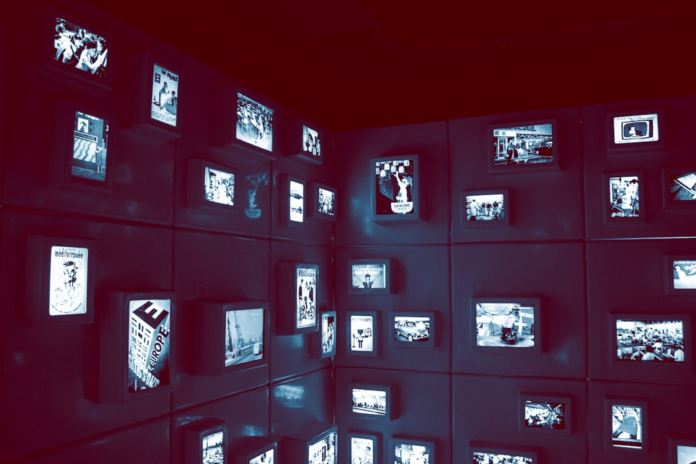 A dark, red-hued wall filled with a grid of glowing digital screens displays diverse images, symbolizing the complex information streams and data visualization of brain-computer interfaces.
