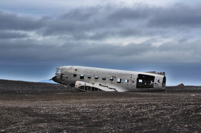 The decaying fuselage of a vintage airplane lies broken on a desolate black sand beach under a heavy, cloudy sky, creating a stark and mysterious scene.