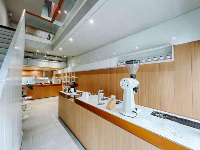 Bright, modern minimalist interior featuring a sleek coffee bar with a prominent white grinder, light wood paneling, white counters, and a visible mezzanine level, conveying a clean and professional environment.