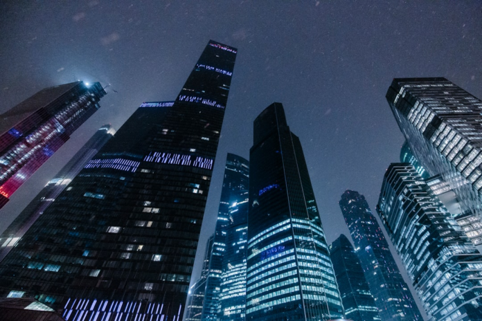 Towering glass-clad skyscrapers with vibrant blue and white lights stand against a dark, expansive night sky, evoking a sense of powerful, futuristic urban complexity.