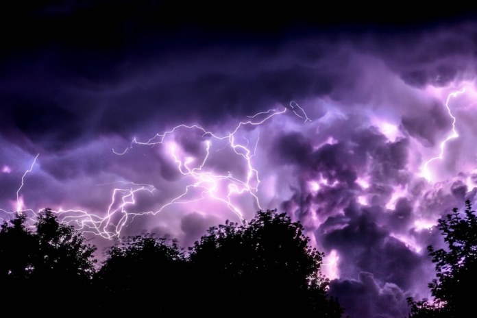 Powerful purple lightning illuminates dark, turbulent storm clouds above silhouetted trees during a dramatic night, symbolizing dynamic atmospheric changes.
