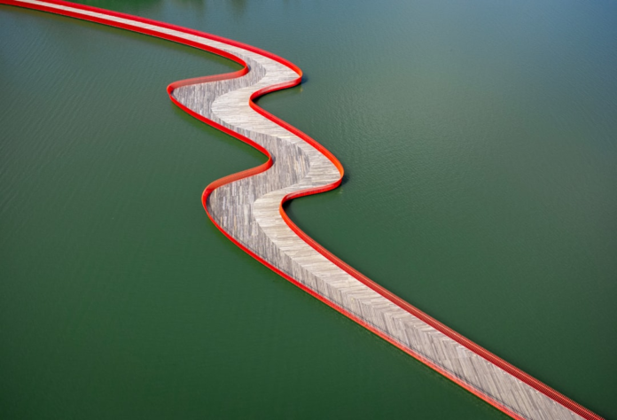 Intricate, undulating wooden bridge with striking red edges gracefully curves across calm dark green water, visually representing the complex and often non-linear paths of human decision-making.