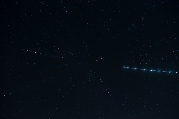 A vast, dark night sky filled with countless stars is crossed by two prominent linear arrays of bright, evenly spaced lights, suggesting active satellite constellations or orbital traffic essential for commercial resupply missions.