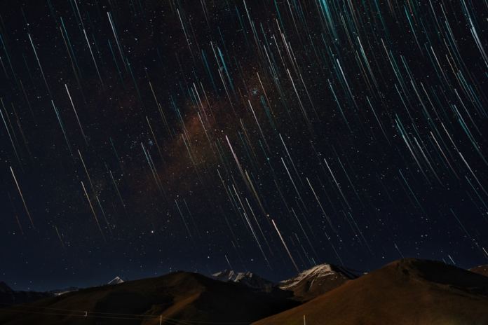 Brilliant star trails streak across a dark, deep blue night sky above silhouetted mountains, conveying the immense scale and timeless journey of cosmic evolution.