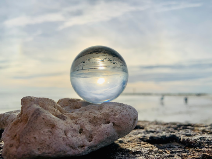 A clear crystal ball, perched on textured rocks, reflects an inverted sun over a tranquil beach scene under a cloudy sky, symbolizing cosmic perspective and understanding.