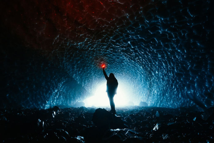A silhouetted figure holding a vibrant red light stands in the middle of a vast, ethereal blue tunnel, glowing brightly from a distant source, evoking the profound mystery of the universe's rapid expansion.