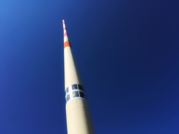 A towering, slender space exploration monument with reflective observation windows and a red-striped pinnacle stretches skyward against a vast, clear blue sky, embodying the ambitious spirit of the Cosmos Program.