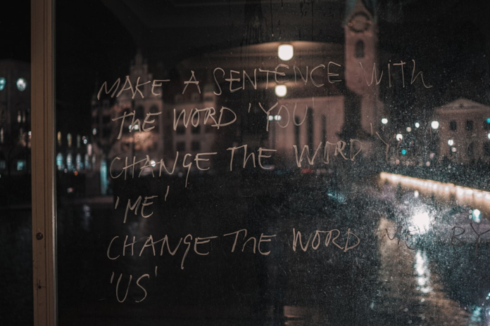 White handwritten text on a smudged windowpane, reading MAKE A SENTENCE WITH THE WORD 'YOU' CHANGE THE WORD 'ME' CHANGE THE WORD 'US', reflects a blurred, illuminated urban cityscape at night, symbolizing evolving human connection and collective identity within society.