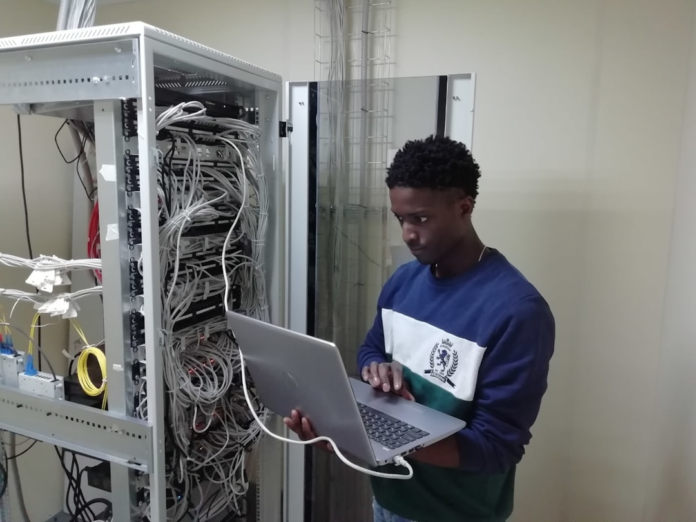 A focused young IT professional uses a laptop while standing next to an open server rack with complex network cabling, representing critical work in data infrastructure and cybersecurity.