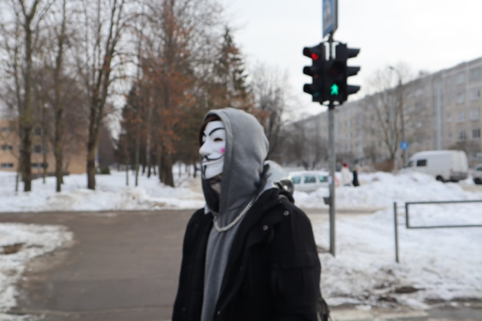An individual wearing a distinctive Guy Fawkes mask and grey hoodie stands on a snowy urban street amidst bare trees and buildings, evoking the anonymous presence of a hacker or cybersecurity 'bad actor'.