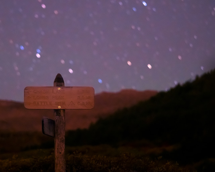 A wooden trail sign points towards distant peaks under a vast night sky filled with numerous blurred stars and silhouetted mountains, evoking the spirit of exploration.