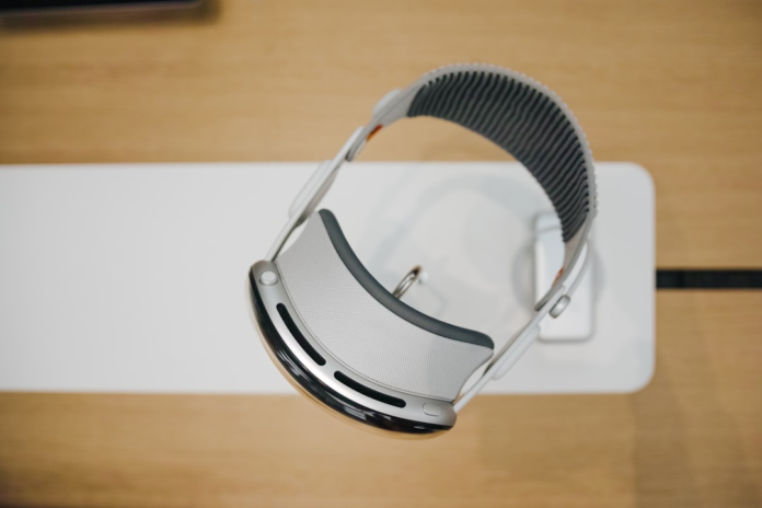A sleek, futuristic silver and gray mixed reality headset, resembling an Apple Vision Pro, is prominently displayed on a clean white stand against a light wooden background, conveying innovation in technology.