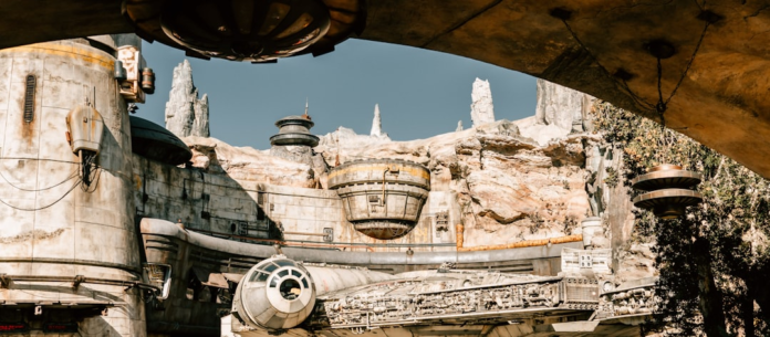 A large, weathered spacecraft resembling the Millennium Falcon is docked in a detailed alien spaceport built into rugged rock formations under a clear sky, evoking a sense of advanced deep space exploration.