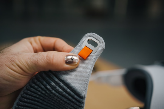 A hand with a metallic thumb nail featuring a star design holds a grey textured strap with an orange pull tab and a silver metal loop, highlighting a tactile connection to digital components.