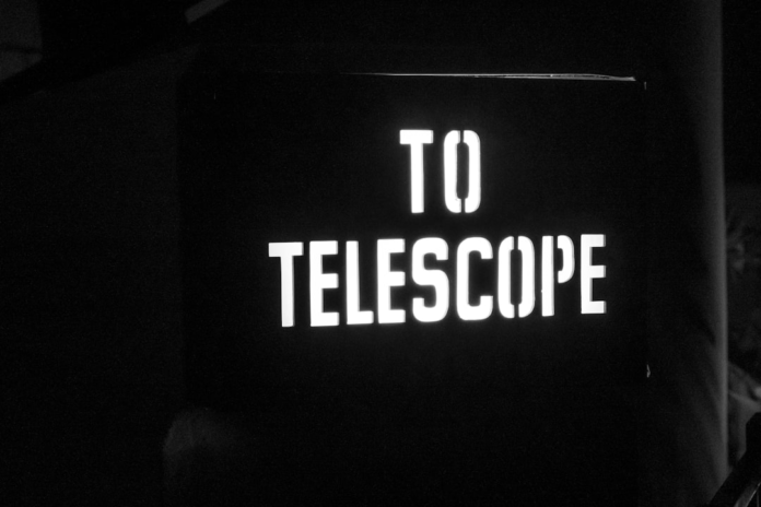 White stencil letters spelling TO TELESCOPE glow against a dark, grainy background, symbolizing the profound search for extraterrestrial life and cosmic discoveries.