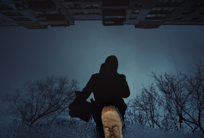 A lone figure's dark silhouette and boot are reflected in a shimmering puddle, capturing an inverted world of distant buildings and bare tree branches under a mysterious, dark sky, evoking a sense of exploration and the otherworldly.