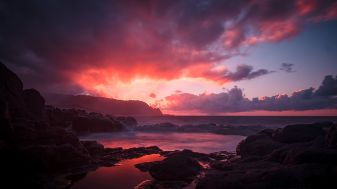 A powerful red and orange sunset fills a cloudy sky above a dark, rugged ocean coastline with smooth water, illustrating atmospheric weather systems.