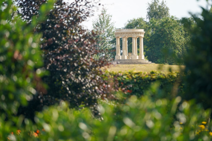 Classical stone rotunda with columns on a sunlit grassy hill, framed by lush green and dark foliage, evoking the sacred tranquility of ancient Greek temple grounds.