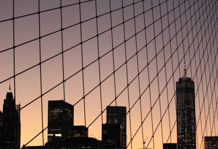 The silhouetted New York City skyline, featuring One World Trade Center, is viewed through the prominent diagonal cables of a bridge against a dramatic twilight sky gradient.