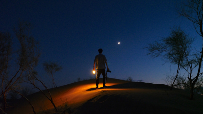 A lone figure holds a glowing lantern on a dark sand dune, looking up at the crescent moon in the vast night sky, symbolizing the profound human search for alien life and answers to the Fermi Paradox.