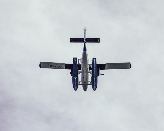 White and blue seaplane viewed from directly below, showcasing its floats and undersides as it soars against a light grey, overcast sky.