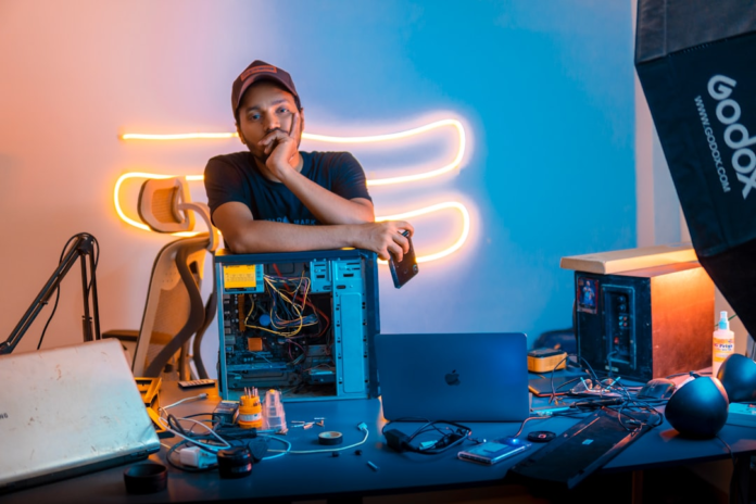 A focused man leans on an open computer tower amidst a desk cluttered with laptops and tech tools, bathed in the vibrant orange and blue glow of a neon-lit digital workspace, embodying hands-on engagement with technology.