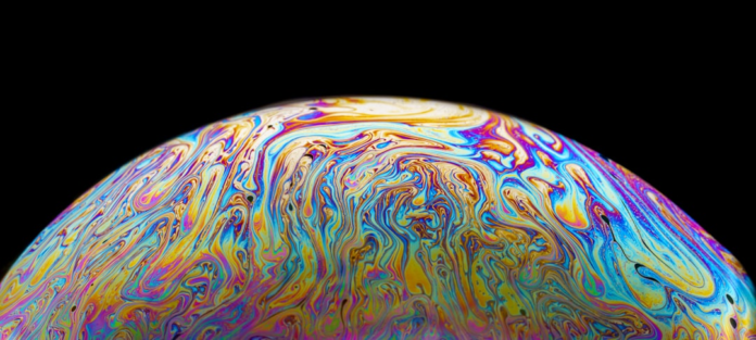 Iridescent blues, purples, and yellows swirl across a curved surface, visualizing the dynamic atmosphere or deep liquid oceans of the waterworld exoplanet GJ 1214 b.