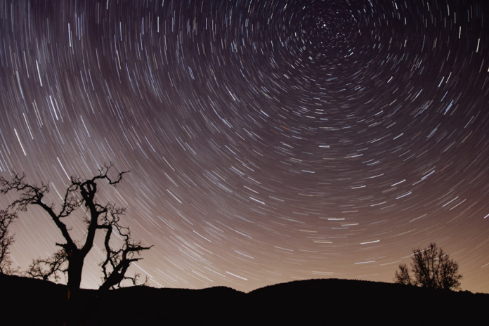 Vast star trails spiral across the dark night sky above silhouetted gnarled trees and distant hills, emphasizing the Earth's rotation and the grandeur of the cosmos.