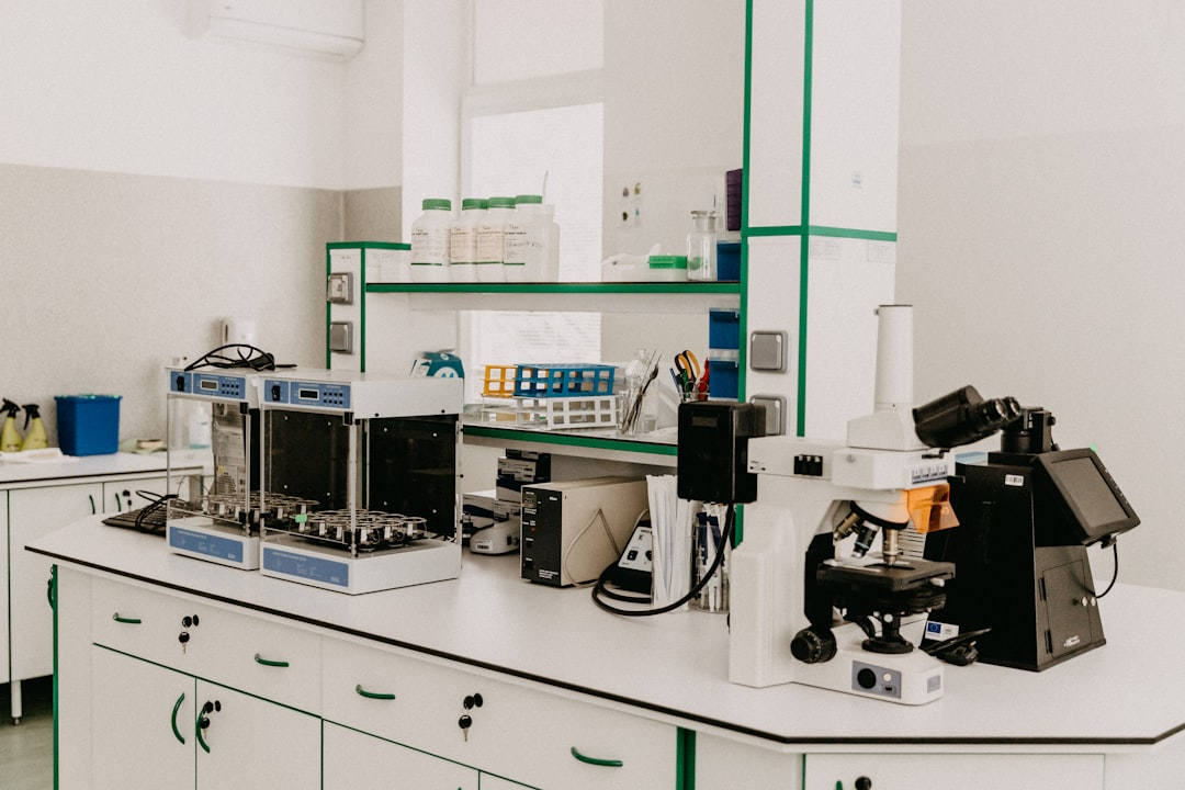 A clean, modern scientific laboratory features advanced microscopes, shakers, and various equipment on white countertops with green accents, suggesting a facility dedicated to genetic engineering and biotechnology research.