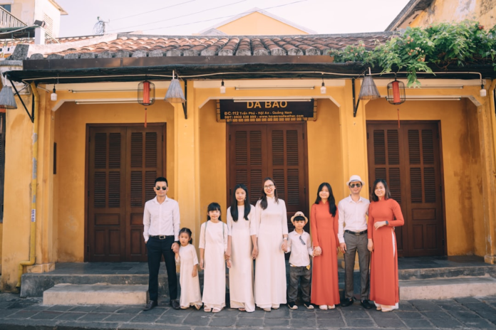 A large, multi-generational Asian family, some in traditional Ao Dai, stands proudly before a warm yellow building in Hoi An, illustrating the concepts of heredity and inherited genetic traits.