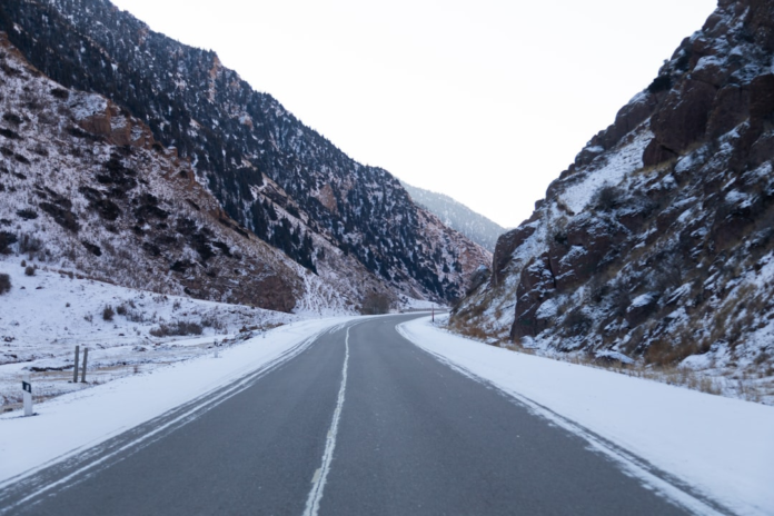 A snowy mountain road curves through a majestic U-shaped valley, flanked by steep, snow-dusted peaks covered with dark evergreen trees, illustrating landscapes shaped by glacial forces.