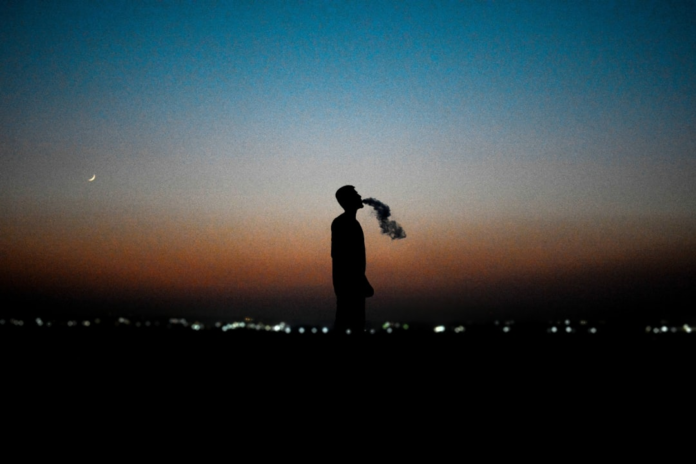 A solitary human silhouette exhales dark vapor against a dramatic twilight sky featuring a crescent moon and distant blurred city lights, evoking contemplation of the unknown or extraterrestrial mysteries.